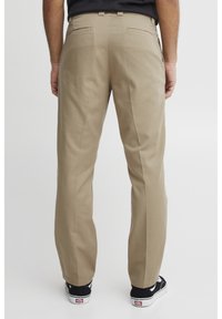 Khaki Chinos mit geradem Schnitt, ausgestattet mit zwei Gesäßtaschen, einer glatten Textur und kombiniert mit schwarzen Sneakers.