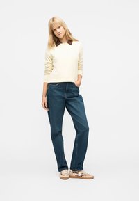 Ξανθιά γυναίκα που φοράει κρεμ πουλόβερ με καφέ γιακά, μπλε τζιν και μπεζ παπούτσια με σχέδιο, στέκεται με το ένα χέρι στην τσέπη μπροστά σε λευκό φόντο.