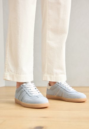 Zapatillas de ante azul claro con cordones blancos y un talón beige, con rayas laterales en contraste y suela de goma, combinadas con pantalones de pana blancos.