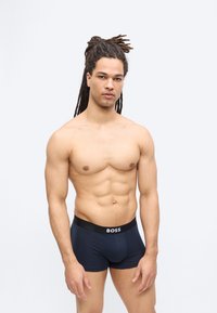 Homme en forme avec des dreadlocks portant un boxer BOSS noir, debout devant un fond blanc uni.