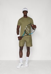 Vihreä oliivinvärinen t-paita ja shortsit, valkoinen rannekoru, valkoinen lippalakki, pitää siniväristä tennismailaa, parina valkoiset tennislenkkarit ja sukat.