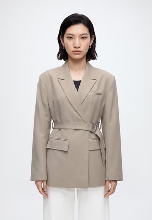 Femme aux cheveux courts et foncés portant un blazer beige ceinturé sur un haut noir et un pantalon blanc, debout devant un fond blanc uni.