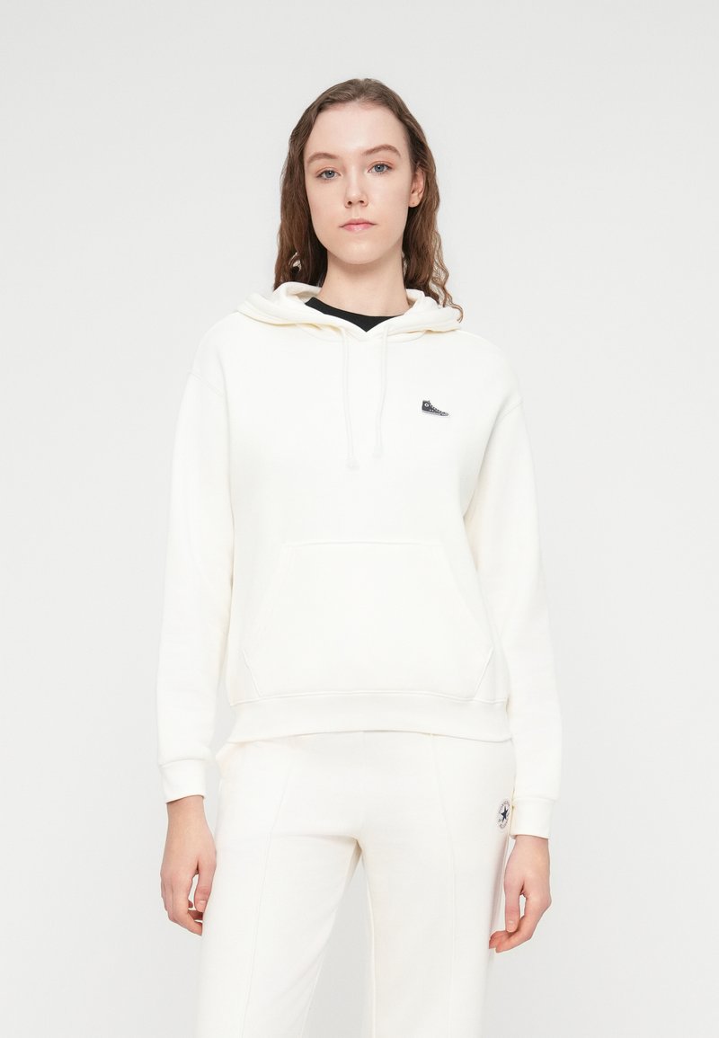 Sweat à capuche blanc en tissu doux, avec une poche frontale, une capuche à cordon de serrage et un petit logo sur la poitrine. Texture lisse, coupe décontractée.