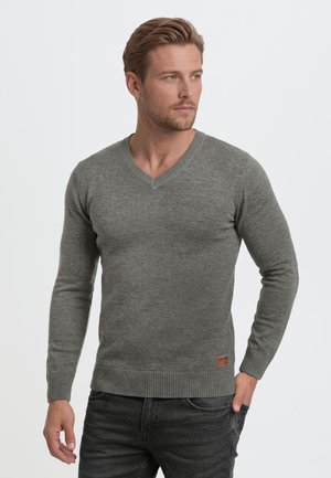 Homme aux cheveux courts châtain clair portant un pull gris à col en V et un jean foncé, debout avec une main dans la poche, regardant sur le côté.