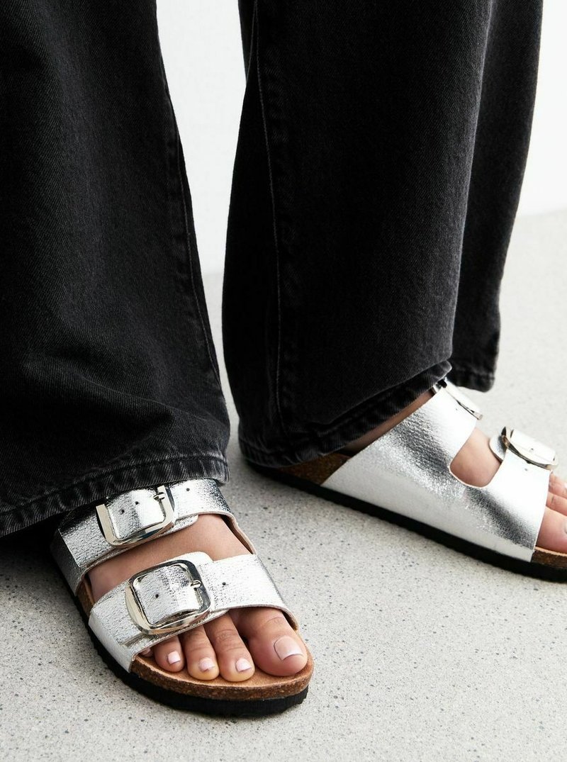 Sandalias metálicas plateadas con dos correas de hebilla, amplias aberturas para los dedos y una suela de corcho. Combinadas con pantalones de mezclilla negros sobre una superficie clara.