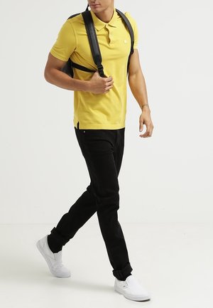 Un homme portant un polo jaune, un pantalon noir, des chaussures blanches sans lacets et un sac à dos noir, marchant devant un fond uni clair.