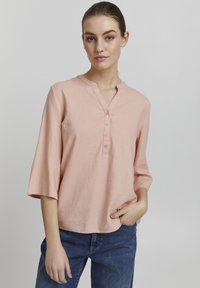 Blouse en lin rose clair avec un col mandarin, patte de boutonnage à cinq boutons et manches trois-quarts, portée avec un pantalon en denim bleu.