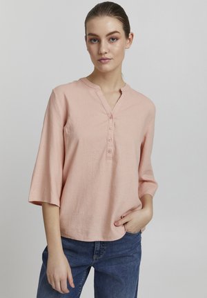 Blouse en lin rose clair avec un col mandarin, patte de boutonnage à cinq boutons et manches trois-quarts, portée avec un pantalon en denim bleu.