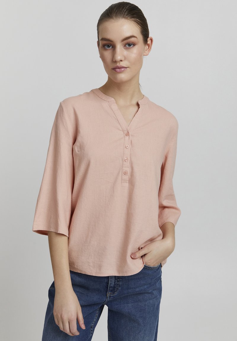 Blouse en lin rose clair avec un col mandarin, patte de boutonnage à cinq boutons et manches trois-quarts, portée avec un pantalon en denim bleu.
