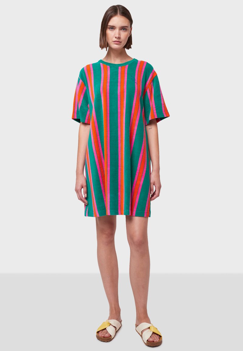 Pletena haljina s vertikalnim prugama u zelenoj, ružičastoj i narančastoj boji. Kratki rukavi, slobodan kroj i okrugli izrez. Uparena s bež i žutim sandalama.