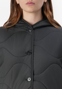 Horná časť trupu modelu zobrazuje detailný záber na čierny prešívaný kabát s kapucňou a dvoma viditeľnými gombíkmi. Vlasy rámujú tvár.
