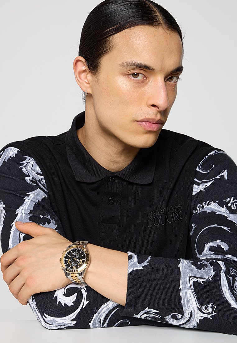Chemise polo noire à manches longues avec un motif grisé tourbillonnant, assortie d'une montre dorée avec un cadran noir et des détails chronographes.