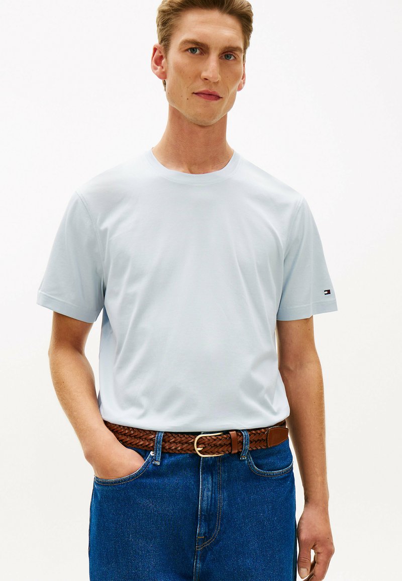 Homme portant une chemise bleu clair à manches courtes rentrée dans un jean bleu avec une ceinture tressée marron, debout avec une main dans la poche.