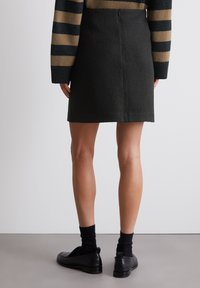 Gonna fitted verde scuro fino al ginocchio con tessuto texturizzato, zip sul retro e orlo diritto. Indossata con scarpe nere e calze.