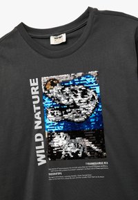Zwarte katoenen t-shirt met een bedrukking van een dinosaurus in pailletten in blauw, zilver en zwart. De tekst luidt "WILD NATURE" met beschrijvingen van soorten eronder.