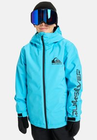 Veste de ski bleue avec fermeture éclair à l'avant, accents noirs et logo. Accompagnée de gants noirs et d'un bonnet, portée avec de grandes lunettes de ski miroir.