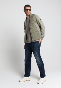 Olijfgroene gewatteerde jas, grijs shirt, donkerblauwe jeans, wit en beige sneakers, zwarte zonnebril, staande houding met handen in de zakken.