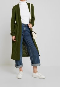 Long gilet vert olive avec un motif en maille texturée, associé à un jean denim bleu taille haute et des baskets blanches. Haut col roulé blanc.