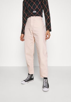 Osoba nosząca jasnoróżowe spodnie z wysokim stanem, czarno-białe wysokie trampki i ciemny sweter w romby z metką "Ragged".