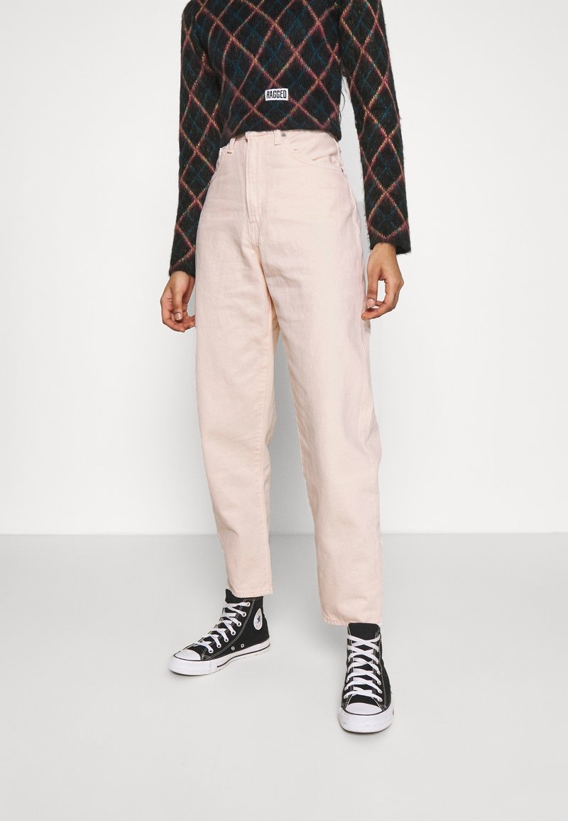 Personne portant un pantalon taille haute rose clair, des baskets montantes noires et blanches, et un pull en losanges foncé avec une étiquette « Ragged ».
