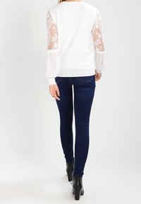 Weißer Pullover mit durchsichtiger Spitze und plissierten Ärmeln, kombiniert mit dunkelblauen Skinny-Jeans und schwarzen Knöchelschuhen.