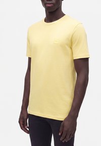 Homme portant un t-shirt jaune uni à manches courtes avec un petit logo carré sur la poitrine et un pantalon foncé, debout devant un fond blanc.