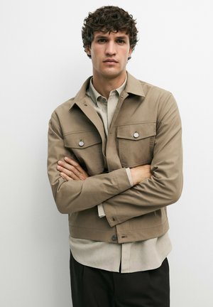 Ung mand med krøllet hår iført en beige jakke over en lys skjorte, stående med armene over kors mod en ensfarvet hvid baggrund.