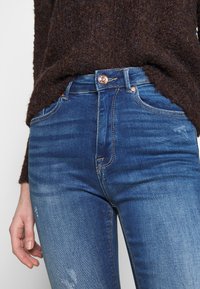 Jeans de denim azul de cintura alta con un acabado ligeramente desgastado, que presentan botones en tono cobre y sutiles detalles de costura.