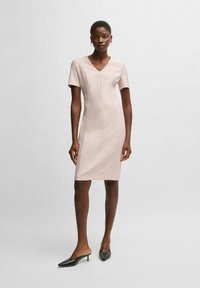 Vestito aderente di un delicato rosa pallido con maniche corte e scollo a V, realizzato in un tessuto morbido. Abbinato a scarpe nere a punta e senza lacci.