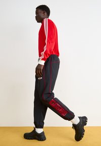 Rød langærmet bluse med hvide striber, sorte bukser med røde accenter og logo, sorte klodsede sko, båret af en person gående i profil.