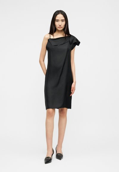 Femme debout portant une robe sans manches en satin noir avec un détail volant asymétrique et des escarpins noirs à bout pointu sur fond blanc.