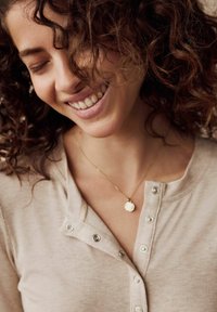 Haut beige à côtes avec des boutons-pression et un collier fin en or avec un pendentif rond. Cheveux bouclés partiellement sur le visage, affichant un sourire.