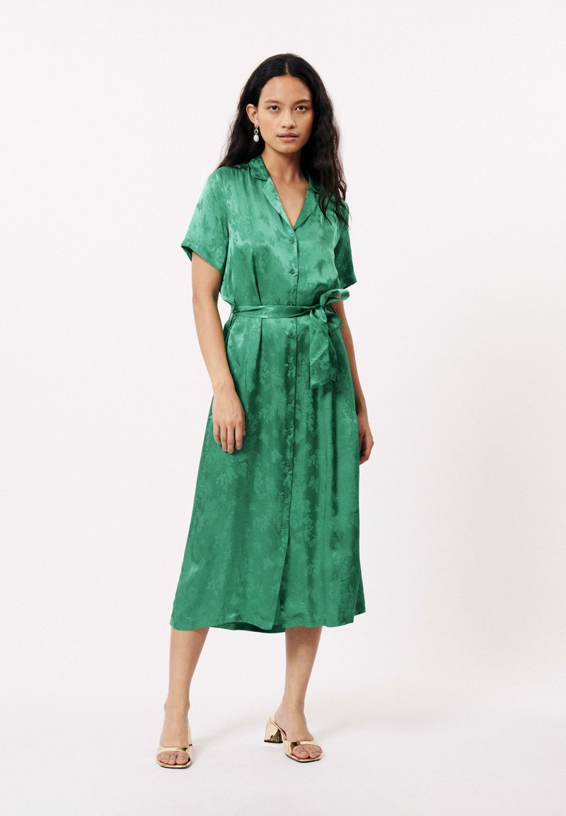 Robe midi en satin vert avec des manches courtes, fermeture à boutons sur le devant, ceinture à nouer à la taille et texture florale subtile. Le modèle porte des sandales avec des accents dorés.