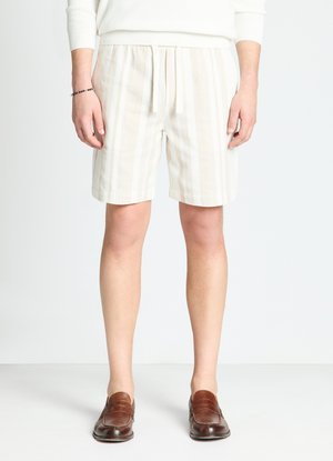 Male wearing white and beige vertical striped shorts with a white knit sweater and brown leather loafers, standing against a plain background.