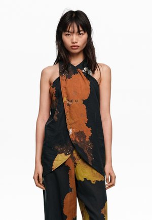 Mujer con cabello largo y oscuro lleva un top sin mangas negro de tipo halter y pantalones a juego con grandes patrones abstractos de salpicaduras de pintura en óxido y amarillo.