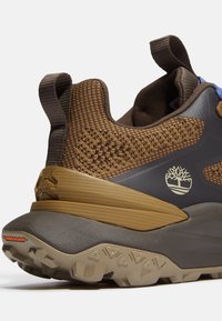 Brown textured sneaker with a gray and tan sole. Features a padded collar, pull tab, and logo accent on the side. Rubber grip outsole.