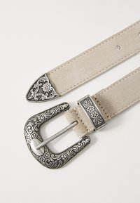 Beige mockaskärp med ett utsmyckat silverfärgat metallspänne med blommönster och spetsig design, som visar på sömmade kanter och justerbara hål.