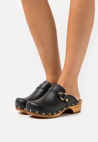 Zwarte lederen klompen met een houten zool, voorzien van een decoratieve messing gesp en studs langs de rand. Gesloten neus en open achterzijde ontwerp.