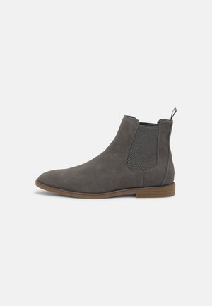Grå ruskind Chelsea ankelstøvle med brun sål, elastisk sidepanel og trækskabelon bagpå på en ensfarvet hvid baggrund.