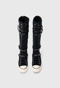 Schwarze knielange Schnürstiefel mit weißen Gummi-Spitzen, ausgestattet mit mehreren schwarzen Riemen und silbernen Schnallen auf einem schlichten weißen Hintergrund.