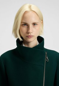 Junge Person mit kurzen blonden Haaren, die einen dunkelgrünen High-Collar-Mantel mit einem silbernen Reißverschluss vor einem schlichten hellen Hintergrund trägt.