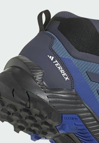 Blå och svarta vandringsskor med texturerad ovandel, förstärkt tå och uttalade sulor. Har 'TERREX'-logotyp och formar sig efter hälen för stöd.