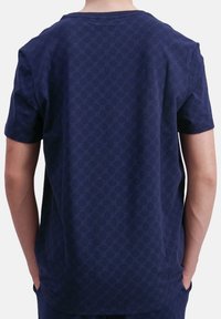 Persoon in een marineblauwe T-shirt met korte mouwen en een subtiel circulair geometrisch patroon, met de handen in de zakken, van zich af gekeerd.
