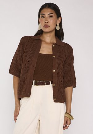 Donna che indossa un cardigan marrone a maniche corte sopra una canottiera abbinata, pantaloni crema a vita alta, orecchini dorati, braccialetti e cintura marrone.