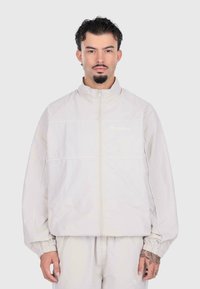 Giacca zip-up grigia chiara con polsini elastici, caratterizzata da cuciture bianche a contrasto. Il modello indossa pantaloni coordinati grigi chiari.