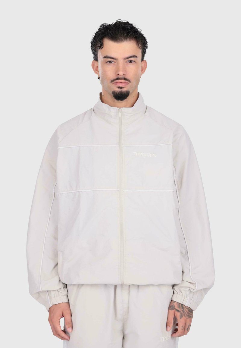 Giacca zip-up grigia chiara con polsini elastici, caratterizzata da cuciture bianche a contrasto. Il modello indossa pantaloni coordinati grigi chiari.