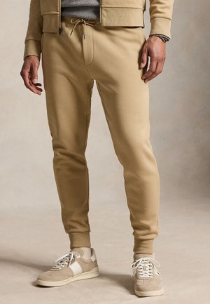 Mężczyzna ubrany w beżowe spodnie dresowe, beżowo-białe tenisówki oraz jasnobrązową bluzę rozpinaną na zamek z szarą koszulką pod spodem.