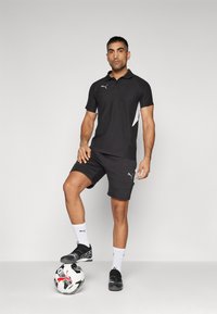 Polo noir avec des accents latéraux blancs et un logo, associé à un short noir. Les chaussettes sont blanches et des chaussures de football noires sont portées. Un ballon de football est en dessous.