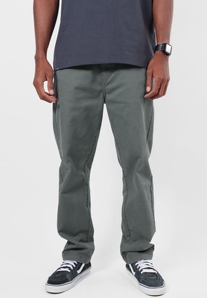 Homme portant un pantalon droit vert foncé, des baskets noires et blanches, une chemise sombre et une montre connectée noire, debout devant un fond uni.