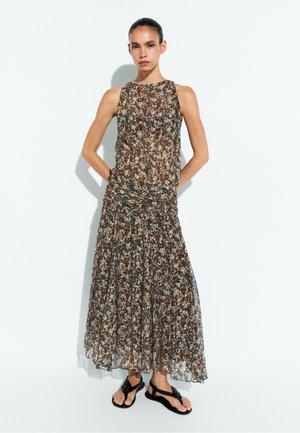 Femme portant une robe maxi marron à fleurs sans manches avec une taille cintrée et des sandales plates noires, debout sur un fond blanc.